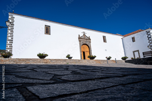 A large white building with a brown door and a cross on the roof. The building is surrounded by a courtyard with a bench and potted plants