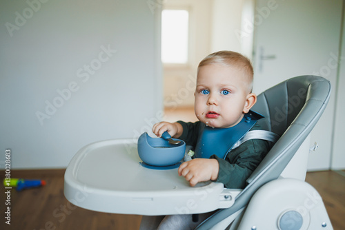 In a bright and minimalist home radiating warmth and joy, a cute little baby sits comfortably in a high chair, wearing an adorable blue bib as they happily enjoy a delightful snack from a bowl