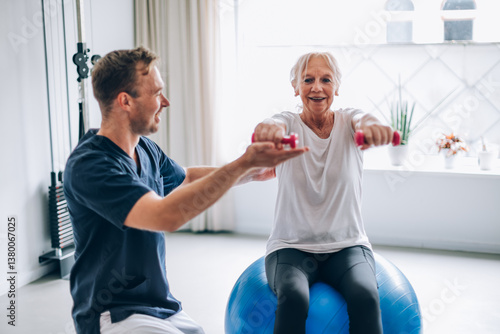 Senior woman lifting weights with physiotherapist on fitness ball: rehabilitation and healthy lifestyle