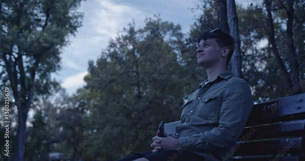 A young man sits on a park bench, holding a vape and looking thoughtfully at the sky. He leans back and takes a deep breath, enjoying the peaceful atmosphere. A young man sits on a park bench, holding a vape and looking thoughtfully at the sky. He leans back and takes a deep breath, enjoying the peaceful atmosphere.