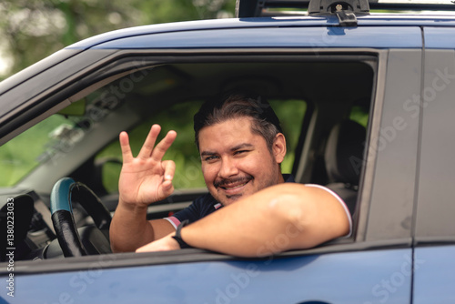 Cheerful middle-aged man smiles and makes an okay hand sign while sitting in a parked car, radiating positivity and assurance.
