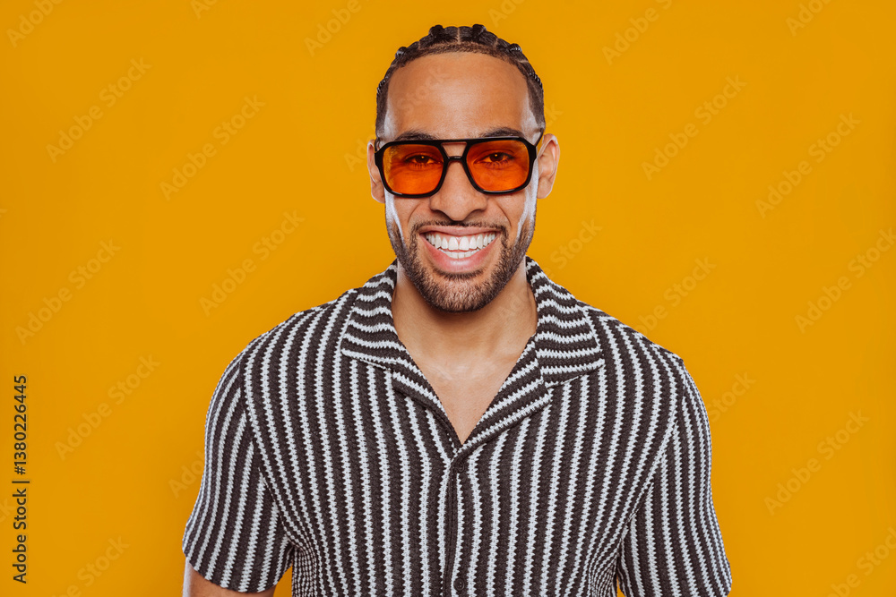 Studio portrait of a cheerful young multiracial man with braided hair, wearing sunglasses and a stylish striped shirt, posing against a vibrant yellow background. Studio portrait of a cheerful young multiracial man with braided hair, wearing sunglasses and a stylish striped shirt, posing against a vibrant yellow background.