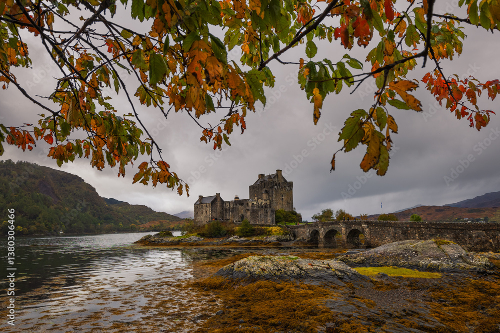 Eilean Donan Castle stands majestically amid autumn hues, its reflection shimmering in calm waters. Misty Highlands and golden trees create a scene of timeless beauty.