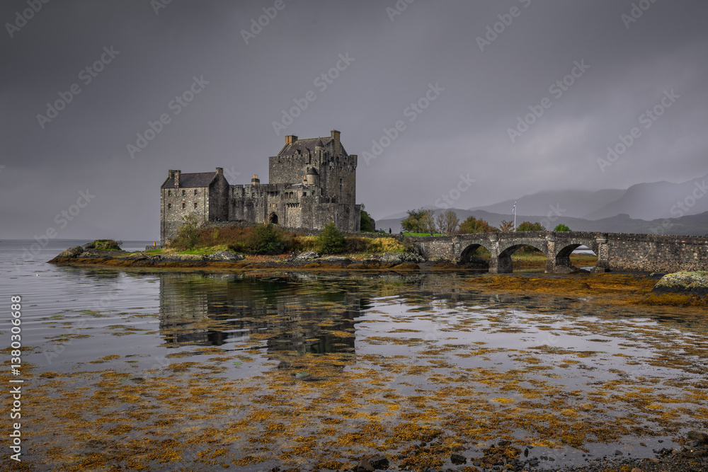 Eilean Donan Castle stands majestically amid autumn hues, its reflection shimmering in calm waters. Misty Highlands and golden trees create a scene of timeless beauty.