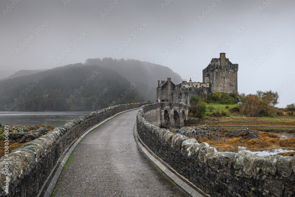 Eilean Donan Castle stands majestically amid autumn hues, its reflection shimmering in calm waters. Misty Highlands and golden trees create a scene of timeless beauty.