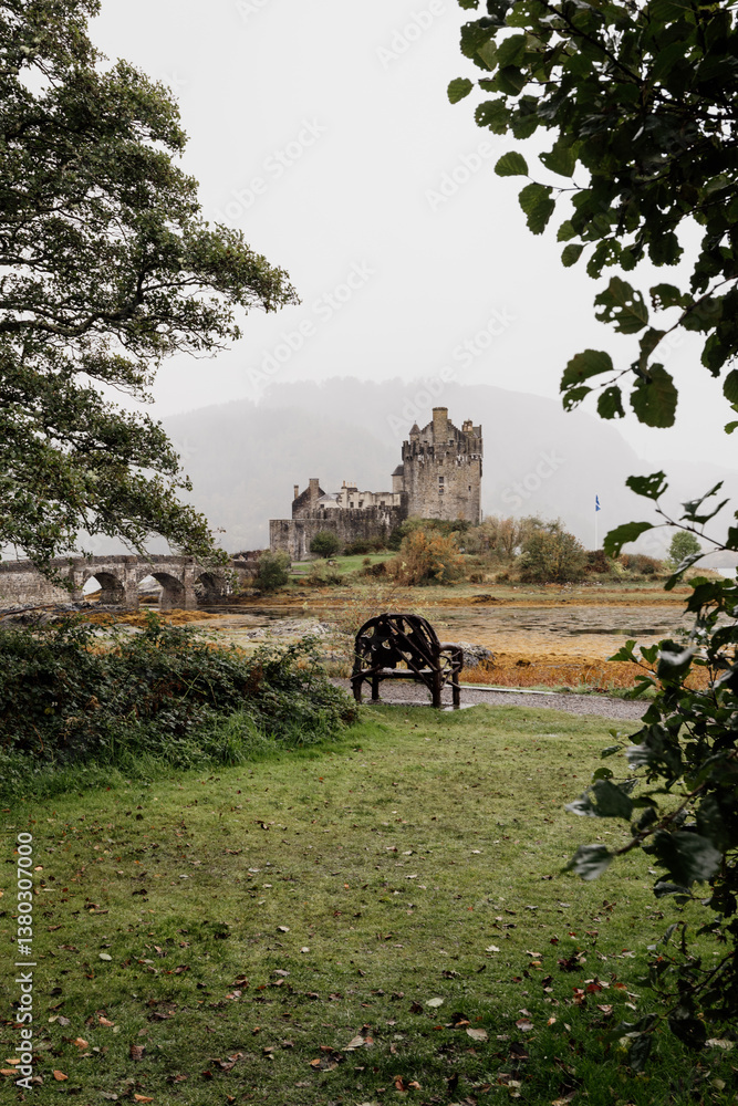Eilean Donan Castle stands majestically amid autumn hues, its reflection shimmering in calm waters. Misty Highlands and golden trees create a scene of timeless beauty.