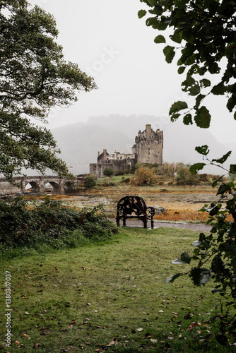 Eilean Donan Castle stands majestically amid autumn hues, its reflection shimmering in calm waters. Misty Highlands and golden trees create a scene of timeless beauty.