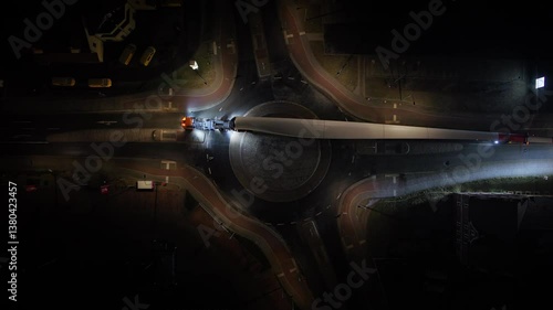 Top-down night shot of a truck transporting a massive wind turbine blade through a roundabout, headlights illuminating the road as it slowly turns right.