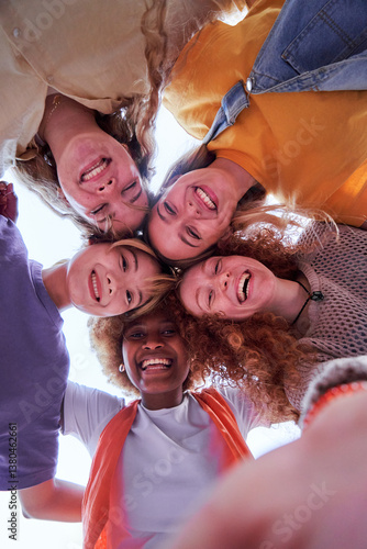 Selfie vertical of multiethnic group of women looking at camera cheerfully embracing each other. Happy female friends in circle looking down. Low angle view. Copy space.