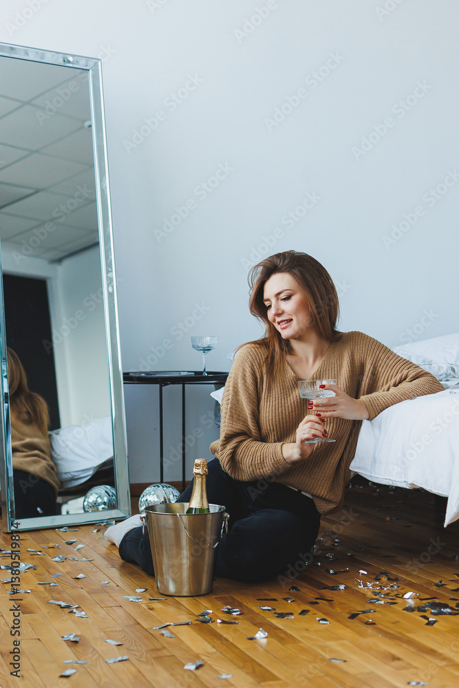 In a stylish bedroom, a young woman enjoys a cozy celebration with champagne, embracing the festive atmosphere with joy, creating lasting memories while relaxing in this charming indoor space