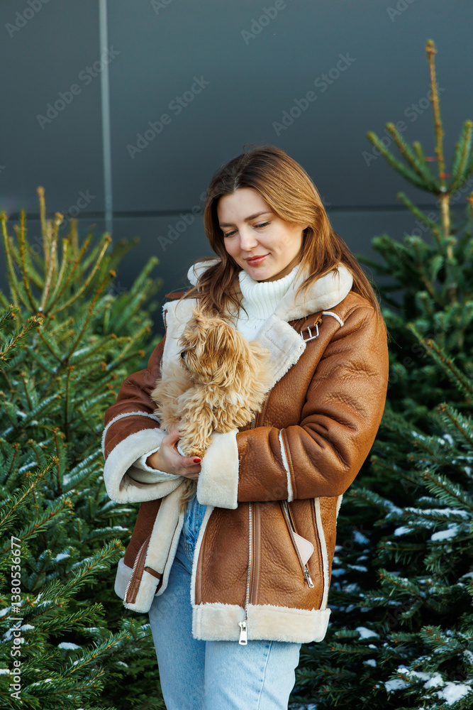 A cheerful young woman warmly embracing her adorable small dog while surrounded by beautifully festive evergreen trees during the enchanting winter season, capturing a perfect moment of joy