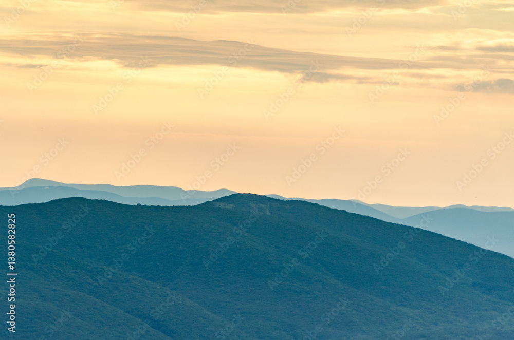 Fototapeta premium Layered mountain ridges under soft, golden dusk light create serene atmosphere. Sky filled with delicate clouds adds tranquility to scene. Calming blend of blue and amber hues