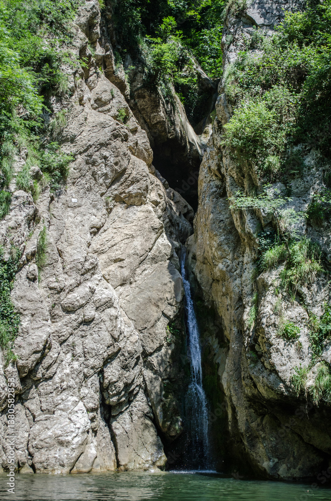Slender waterfall descends through rocky gorge surrounded by lush greenery. Sunlight illuminates scene, highlighting texture of rocks. Water pools at base, forming serene reflections Slender waterfall descends through rocky gorge surrounded by lush greenery. Sunlight illuminates scene, highlighting texture of rocks. Water pools at base, forming serene reflections