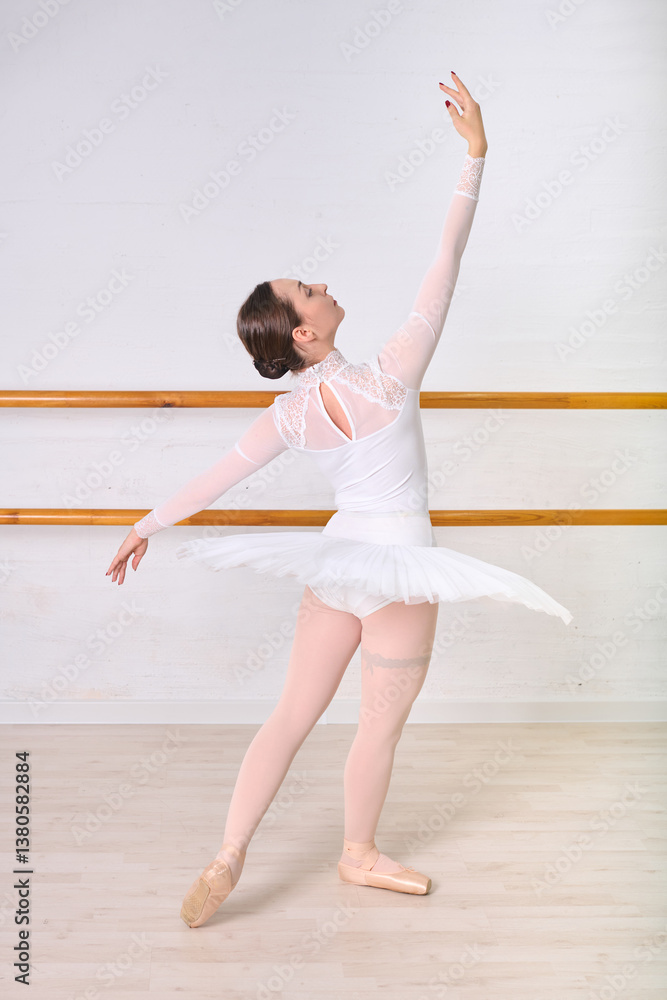 Fototapeta premium Young female ballerina with brunette hair elegantly posing in ballet studio. Dressed in white leotard, embracing classic form against backdrop of wooden barre and light walls