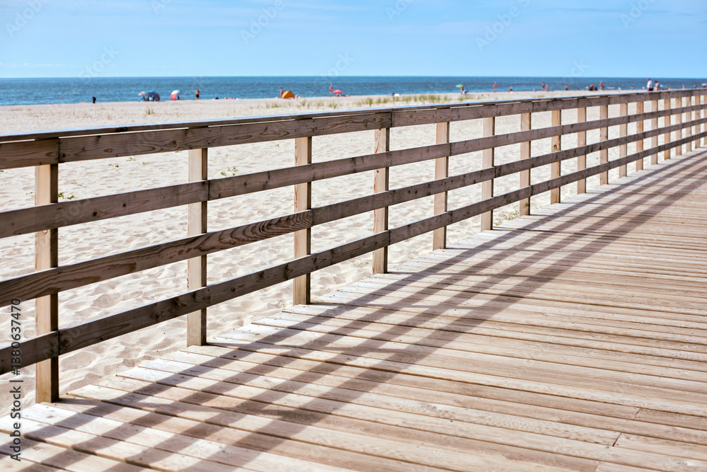 Wooden boardwalk casts shadows across its planks as it meanders beside peaceful sandy beach. In distance, colorful umbrellas dot shore, inviting tranquil seaside escape Wooden boardwalk casts shadows across its planks as it meanders beside peaceful sandy beach. In distance, colorful umbrellas dot shore, inviting tranquil seaside escape