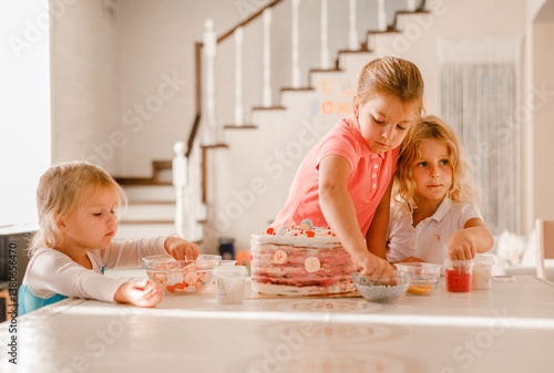 Group of cheerful kids decorating birthday cake together in bright home kitchen during celebration