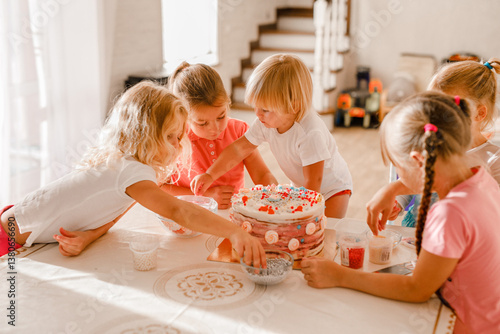 Group of cheerful kids decorating birthday cake together in bright home kitchen during celebration