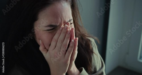 A brunette girl holds her hands to her face and cries near a gray wall at home
