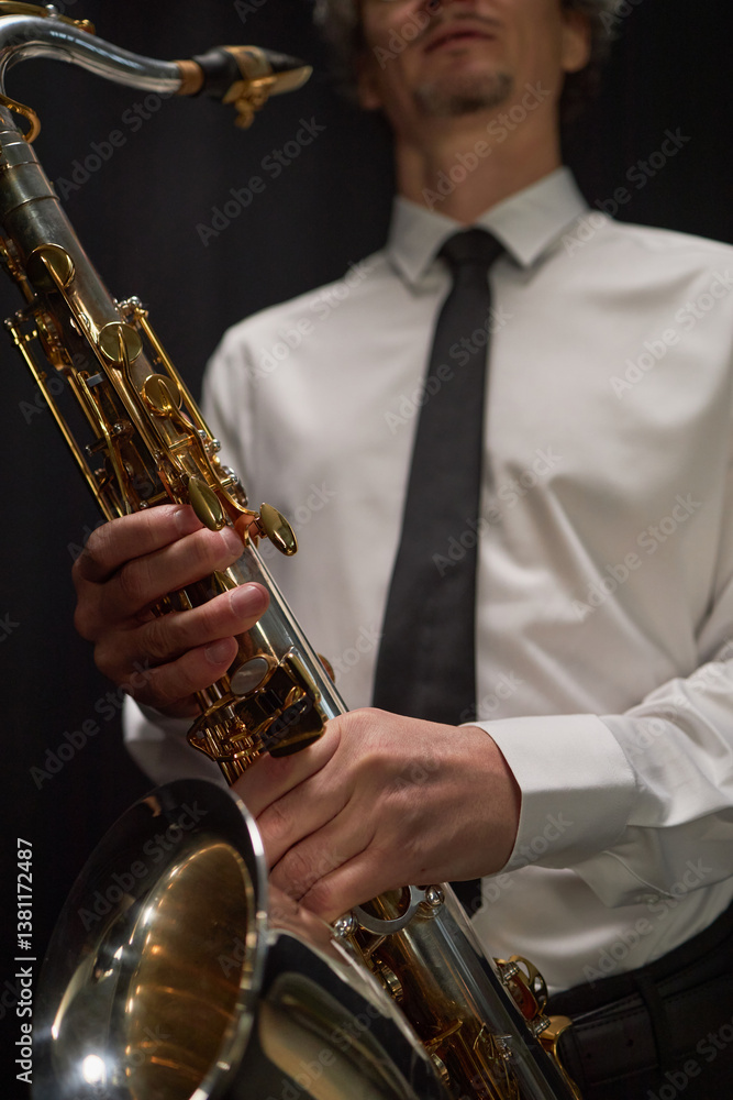Fototapeta premium Vertical cropped shot of professional male musician dressed in formal white shirt and black tie holding tenor saxophone in record studio against black background