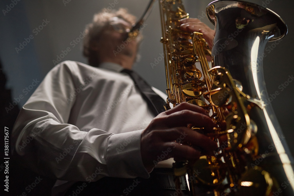 Obraz premium Low angle shot of professional male musician pushing keys of saxophone creating enchanting jazz music with nimble fingers in studio with soft lights