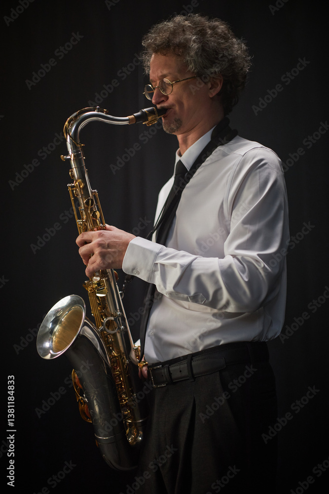 Fototapeta premium Vertical shot of adult male saxophonist dressed in white shirt passionately playing saxophone against black curtains in studio