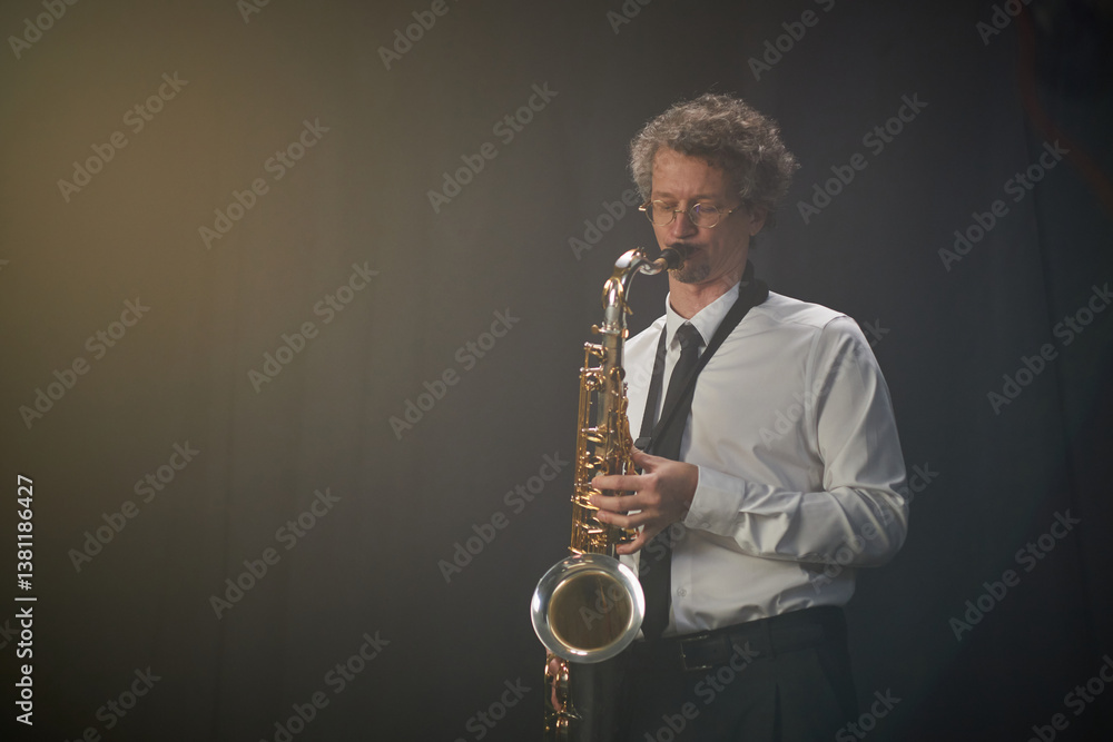Fototapeta premium Adult male saxophonist wearing white shirt and tie playing saxophone with passion standing against backdrop of black curtains in studio with soft lighting