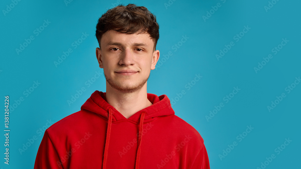Young man in red hoodie with calm expression and soft smile posing against blue studio background. Concept of relaxation, fashion, personality, casual lifestyle routine, peaceful mood, daily vibes. Young man in red hoodie with calm expression and soft smile posing against blue studio background. Concept of relaxation, fashion, personality, casual lifestyle routine, peaceful mood, daily vibes.