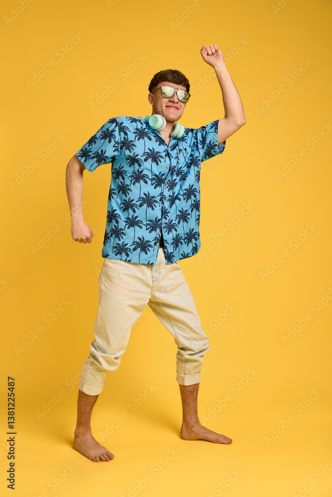 Fototapeta premium Full body photo of expressive young man dancing barefoot with headphones on neck, enjoying holiday moment. Concept of joy, music, self-expression, tropical energy, carefree lifestyle.