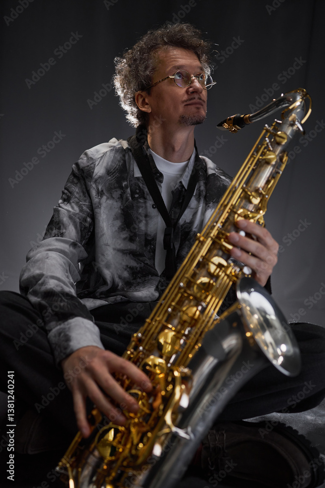 Naklejka premium Vertical portrait shot of middle aged male musician wearing grey shirt and glasses holding saxophone in hands, while seeking inspiration for jazz improvisation sitting in studio with backlight