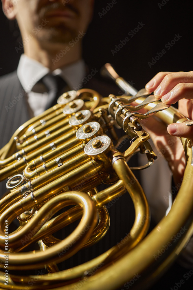 Obraz premium Vertical close up on fingers of male musician pushing valve levers of shiny French horn made of solid yellow brass, while performing deep mellow music on stage under bright spotlight