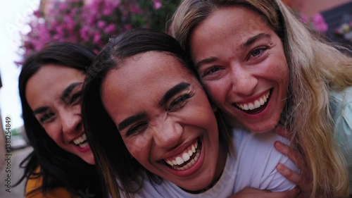Three cheerful female friends are enjoying their vacation together, standing close and looking camera with joyful expressions in a picturesque street adorned with pink flowers