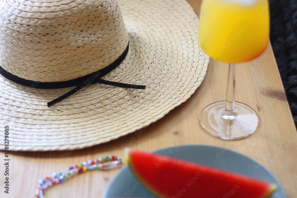 Plate with watermelon slices, glass of orange juice or cocktail, straw hat, colorful beaded necklace and sunglasses on the wooden table. Summer essentials concept. Selective focus.