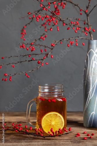 Barberry tea with lemon in a glass jar cup, branches with berries on a wooden table.