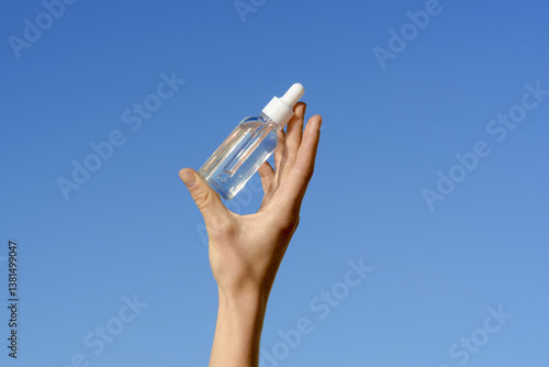 Female hand holding a transparent glass jar of serum with a pipette dispenser on a blue sky background. Concept of beauty and aesthetics products, cosmetics for moisturizing the skin