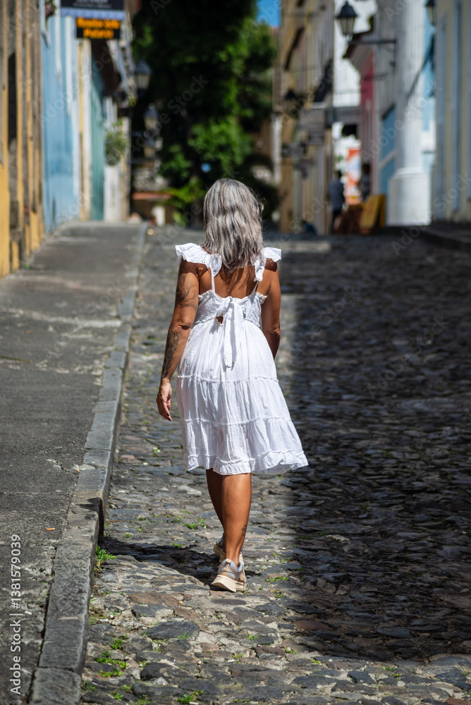 Naklejka premium Beautiful woman wearing white dress and sunglasses, smiling and happy, walking on the streets against old houses. person on travel. South America.