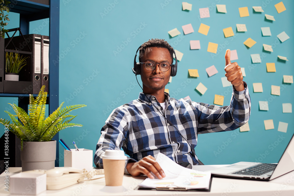 Customer support agent wearing a headset and giving a thumbs up, seated at desk reviewing management charts. African american manager doing an approval gesture towards camera, agreeing with clients.