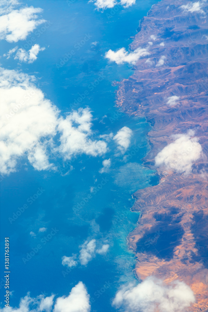 High altitude view of a coastline, showcasing the interplay of land and sea. Aerial view of a striking coastline, where deep blue ocean meets rugged mountains, partially veiled by scattered clouds