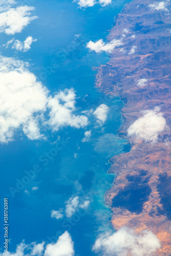 Obraz na płótnie High altitude view of a coastline, showcasing the interplay of land and sea. Aerial view of a striking coastline, where deep blue ocean meets rugged mountains, partially veiled by scattered clouds w salonie