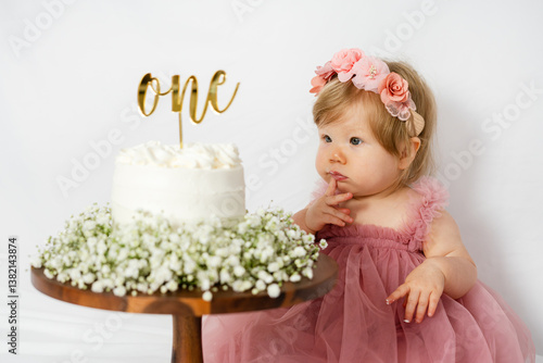 Cute Caucasian baby girl looking at her smash cake on her first birthday. Baby girl birthday cake. One year old birthday celebration. Baby thinking. First cake. 