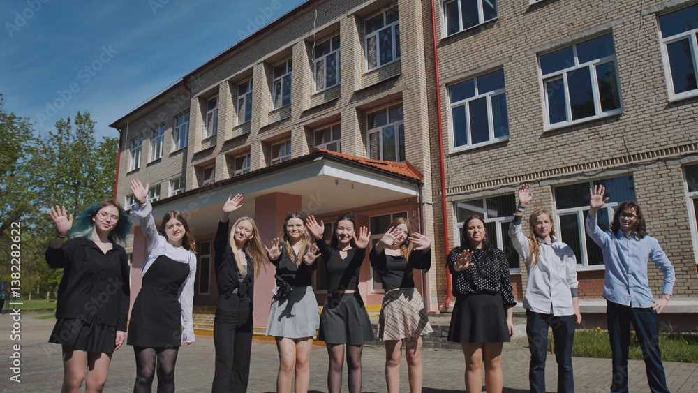 Obraz premium Joyful group of students waving in front of a school building on a sunny summer day, celebrating togetherness and success