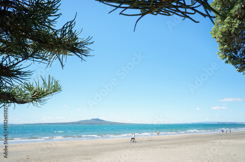 Obraz na płótnie Rangitoto Island framed by Pohutukawa and pine trees. Unrecognizable people walking on the beach. Takapuna Beach. Auckland. w salonie
