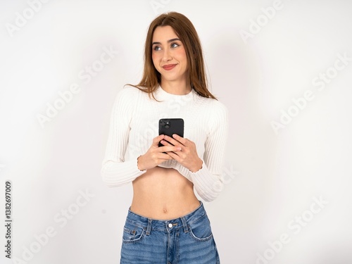 Caucasian young girl wearing white T-shirt holding in hands mobile phone and looking aside with dreamy look.