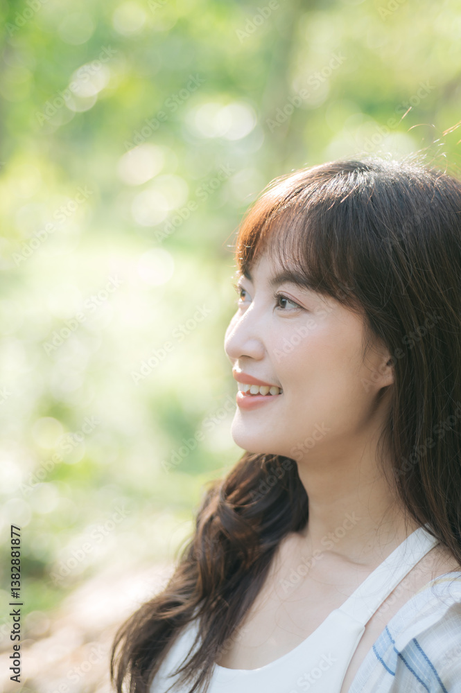 young South East Asian woman with long hair smiles softly while standing outdoors in sunlit, green natural setting. Wellness lifestyle concept. young South East Asian woman with long hair smiles softly while standing outdoors in sunlit, green natural setting. Wellness lifestyle concept.