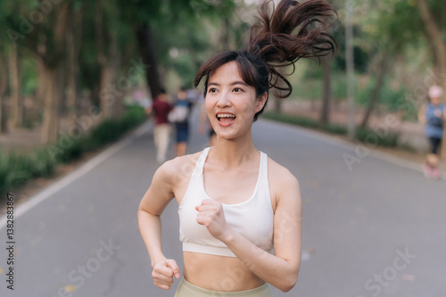 Valokuva Asian woman jogging outdoors in park, wearing white sports bra and smiling with joy, her hair bouncing as she runs on tree lined path