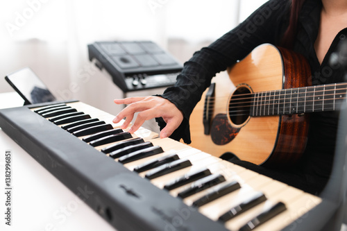 Beautiful Asian teen girl playing guitar, composing music in home recording studio using computer, headphones, condenser microphone, tablet and keyboard for mixing and mastering music.