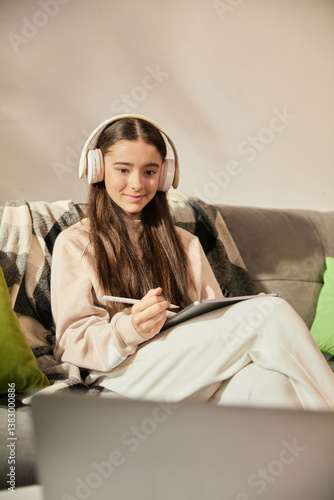 Obraz na płótnie Smiling teen girl sitting on couch in headphones, attending online lesson at home, making notes on digital tablet with stylus. Interactive remote learning at home. Concept of education w salonie