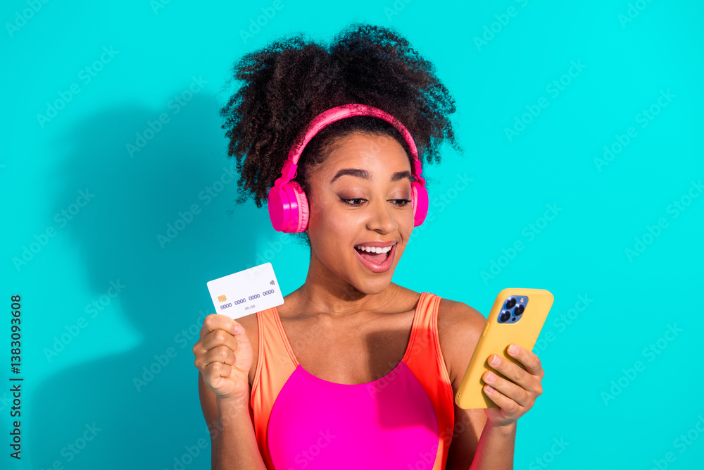 Young woman in vibrant activewear enjoying music with headphones while holding a credit card and smartphone against a teal background Young woman in vibrant activewear enjoying music with headphones while holding a credit card and smartphone against a teal background
