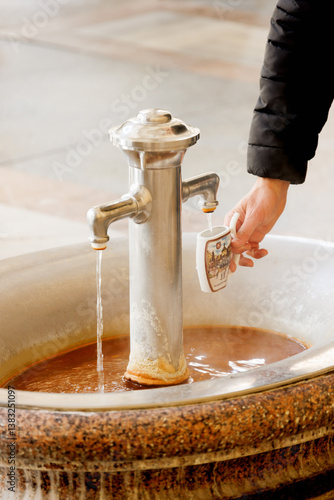 Traditioneller Keramikbecher wird an einer heißen Thermalquelle mit Heilwasser am historischen Kurort Karlovy Vary gefüllt - Karlsbad, Tschechien 2025-03-27