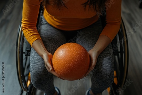 a person is sitting in a wheelchair holding an orange ball in hands, indoors. The person is wearing gray athletic pants and a orange top.