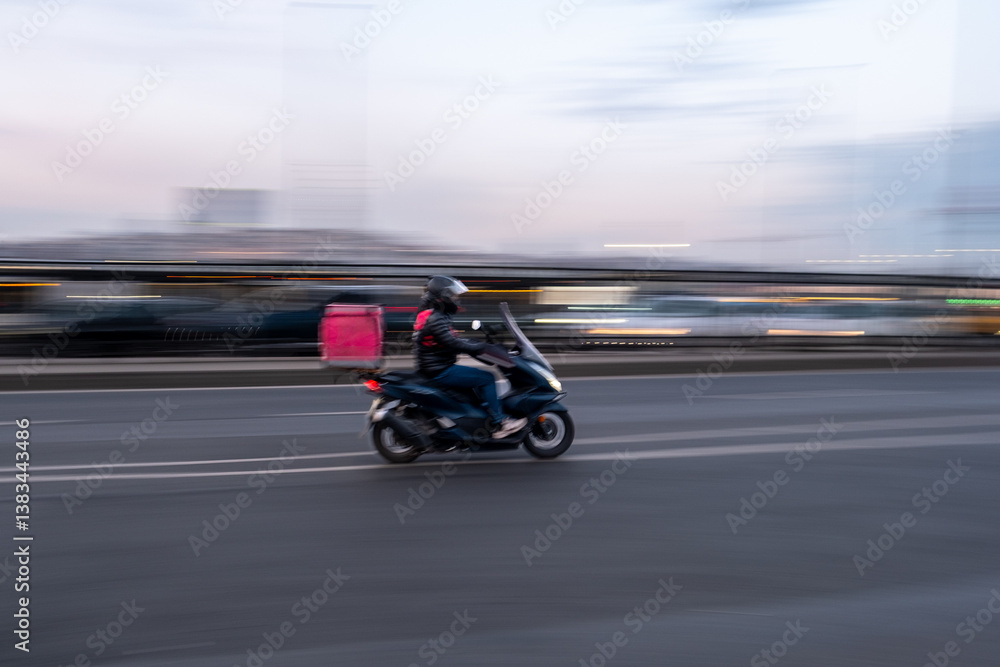 Obraz premium Urban Fast Food Delivery Person on Scooter Racing Through Streets at Dusk: Motion Blur Captures Speed, Efficiency, and Dynamic City Transportation